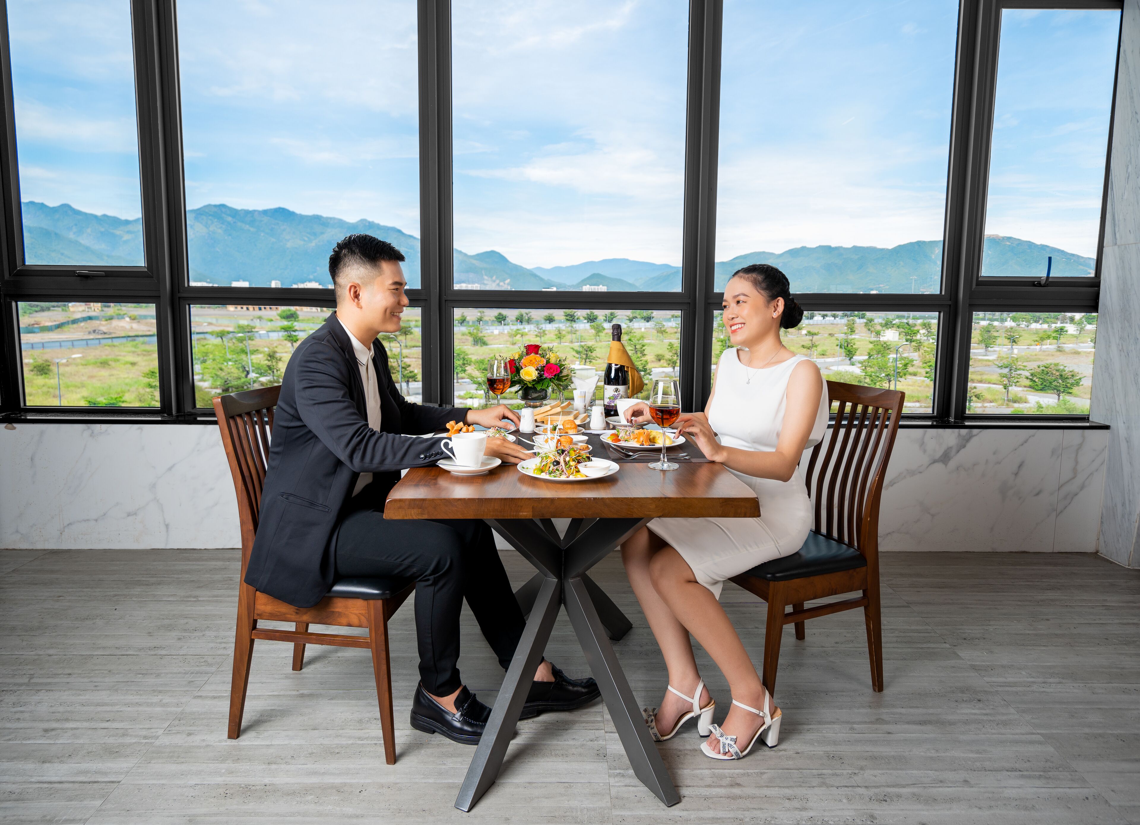 Ein Esstisch mit Blick auf Berge durch große Fenster, zwei Personen sitzen am Tisch, und ein Essen mit Wein.