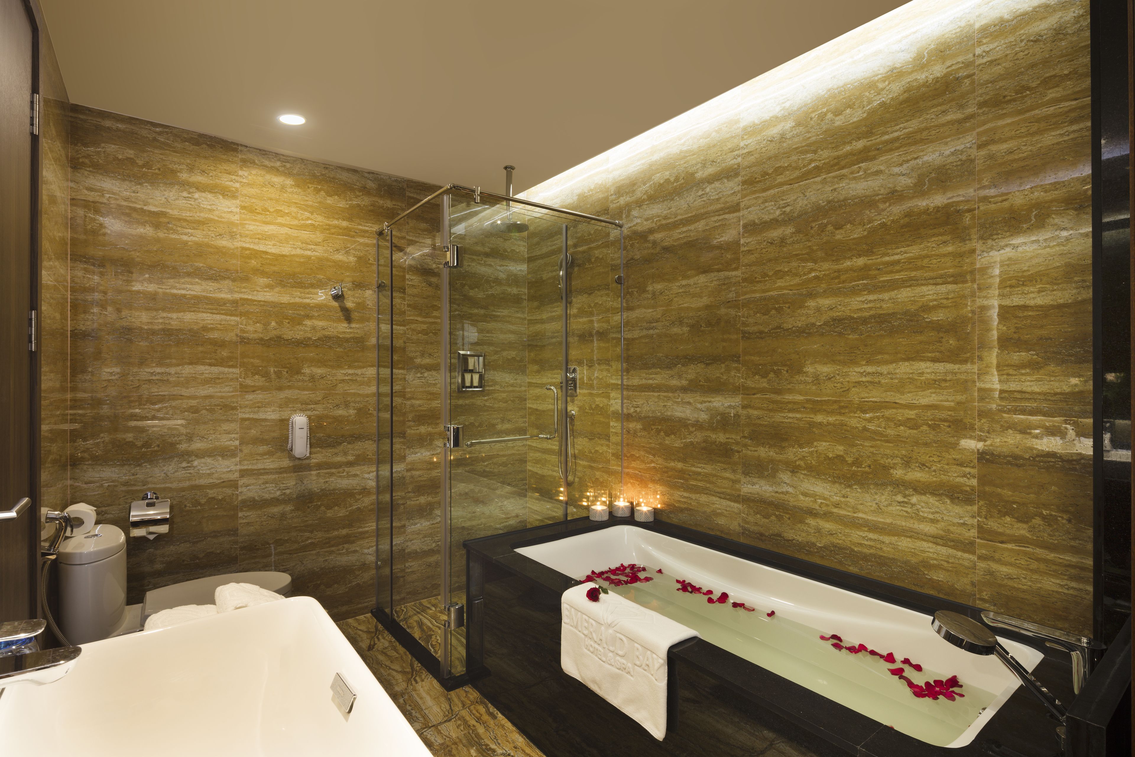 A modern bathroom with a glass-enclosed shower, a large bathtub with rose petals, and a marble wall.