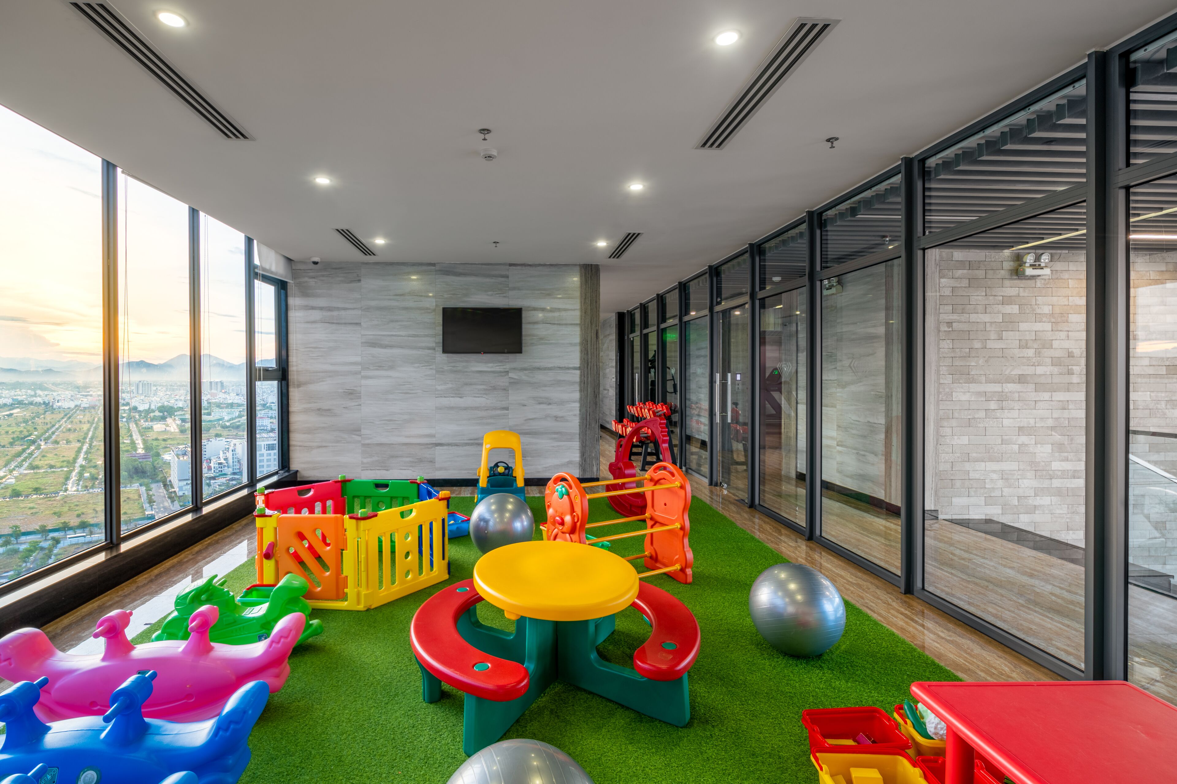 Un espace de jeux pour enfants avec des jouets colorés et une vue sur la ville grâce à de grandes fenêtres.