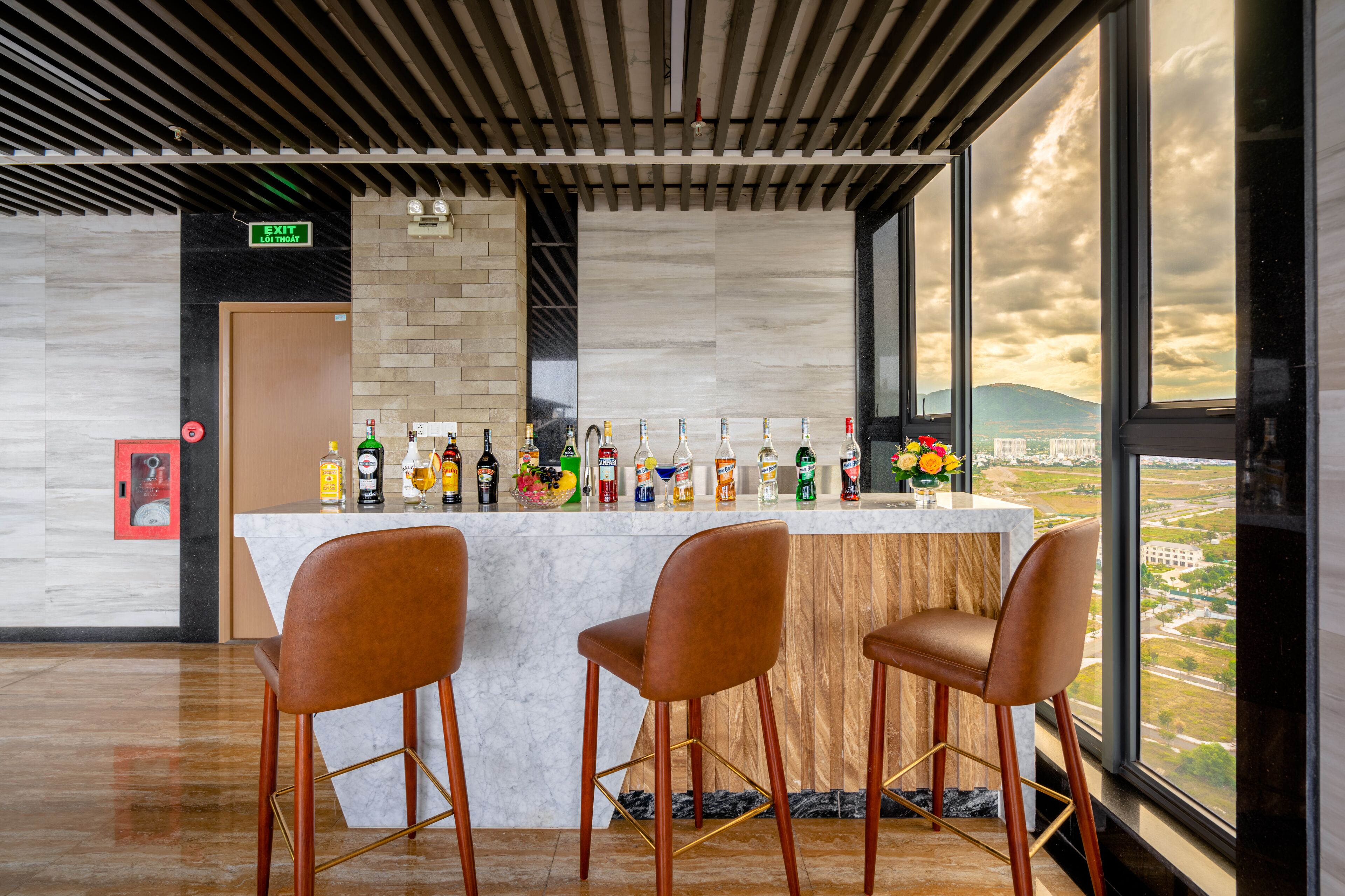 A bar area with a marble countertop, three brown chairs, and a view of the cityscape through large windows.