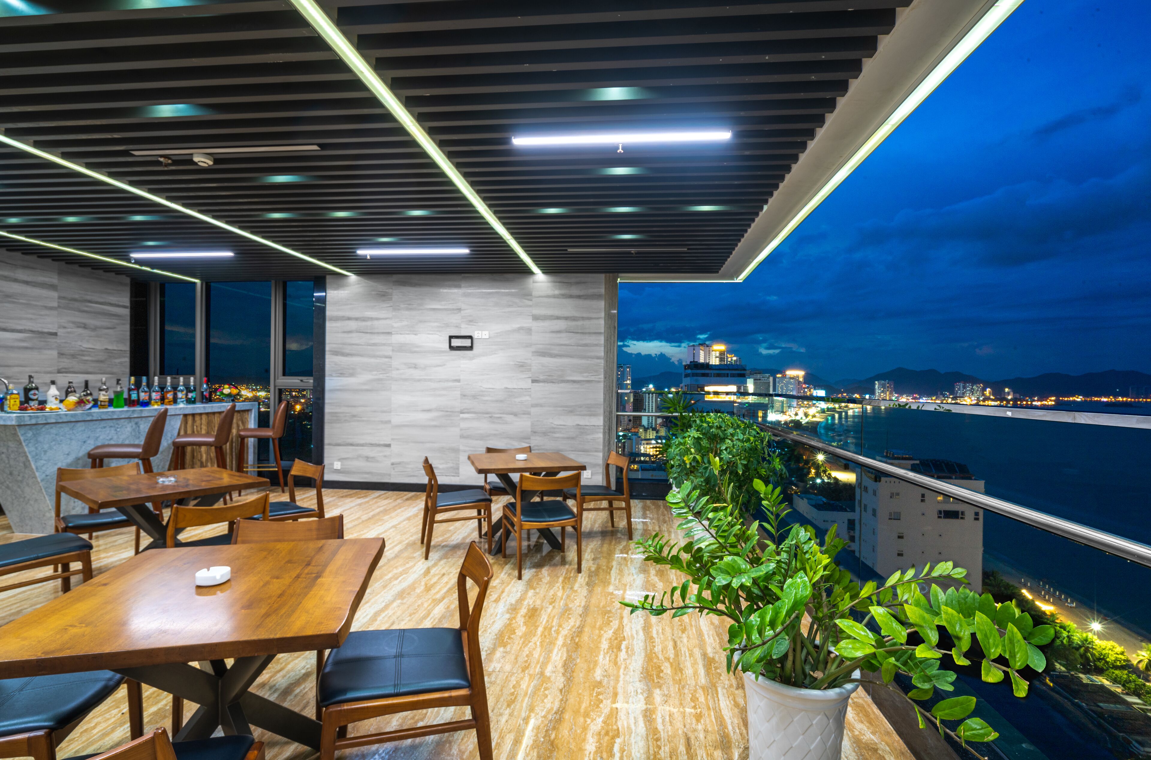 Un restaurant moderne avec des tables et des chaises en bois, un bar et une vue sur la ville et l’eau, de nuit.