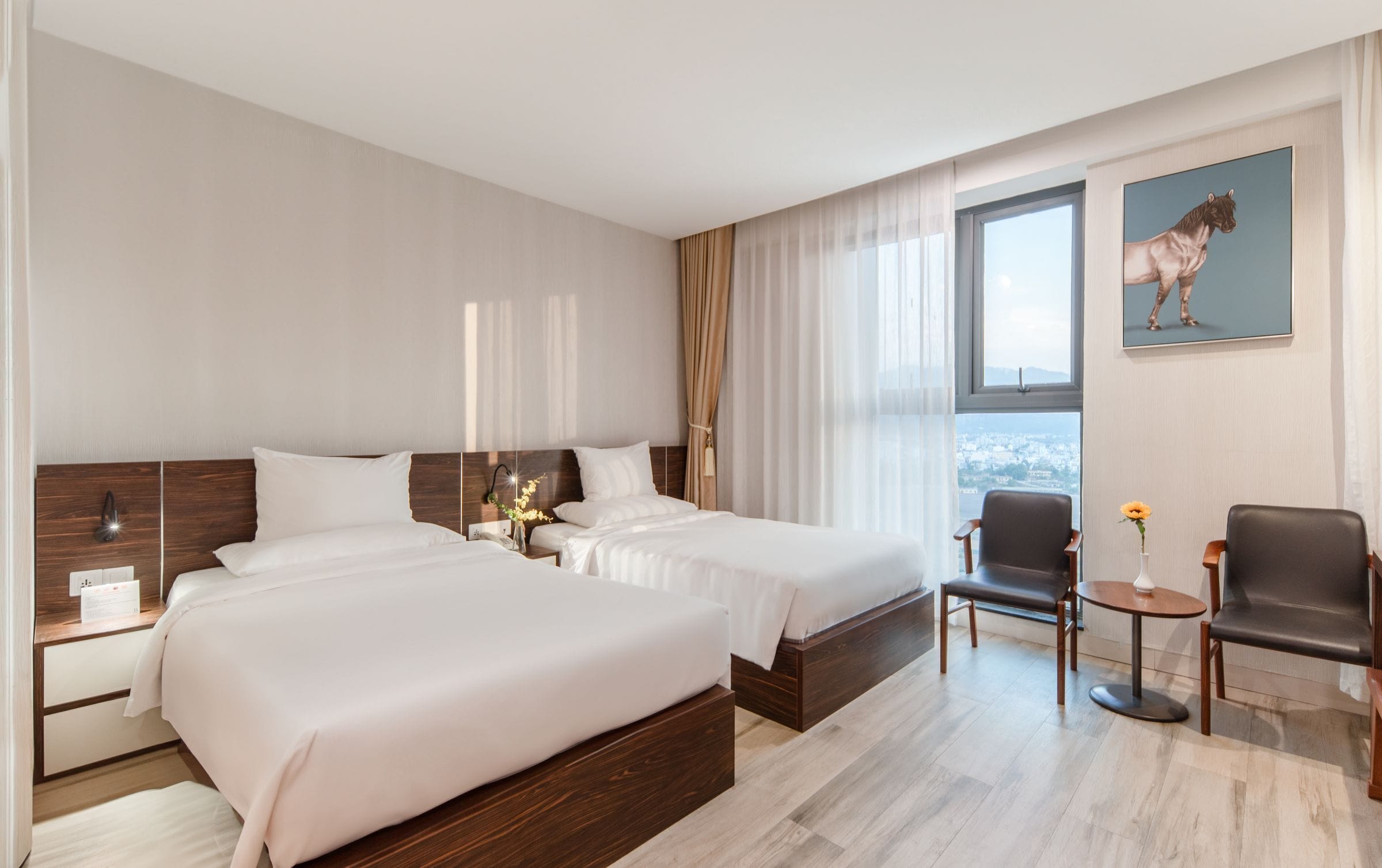 A hotel room with two beds, a desk, a chair, and a window with a city view.