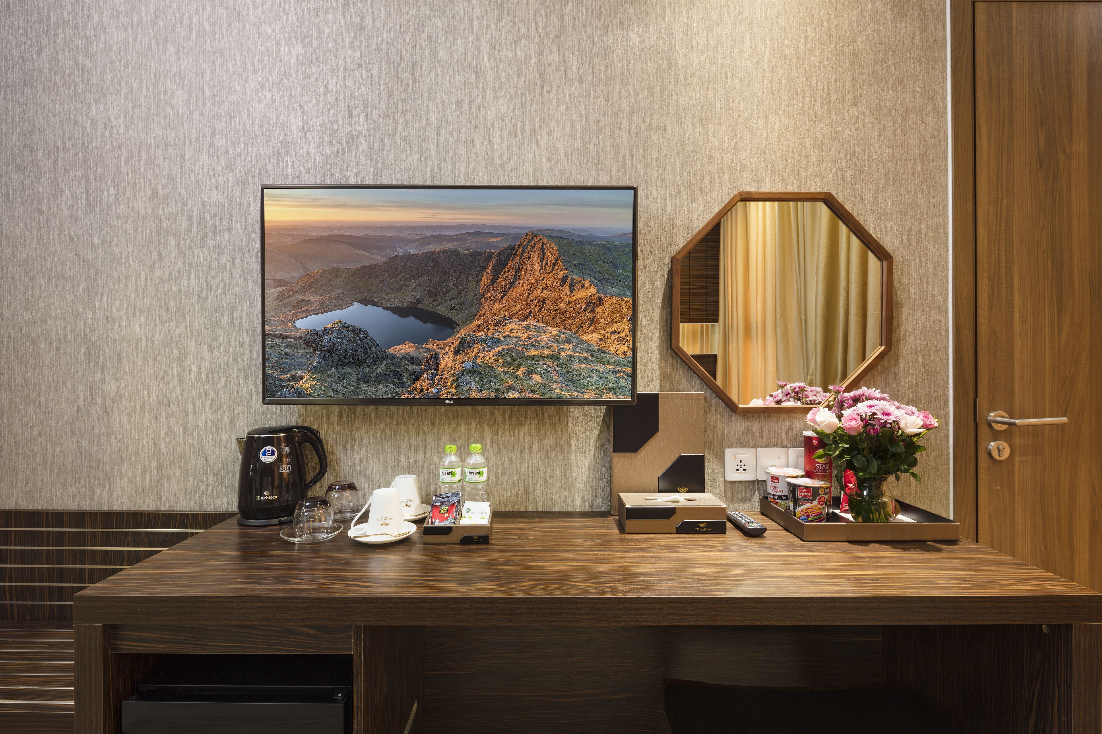 A hotel room with a wooden desk, a flat-screen TV, a mirror, and a vase with flowers.