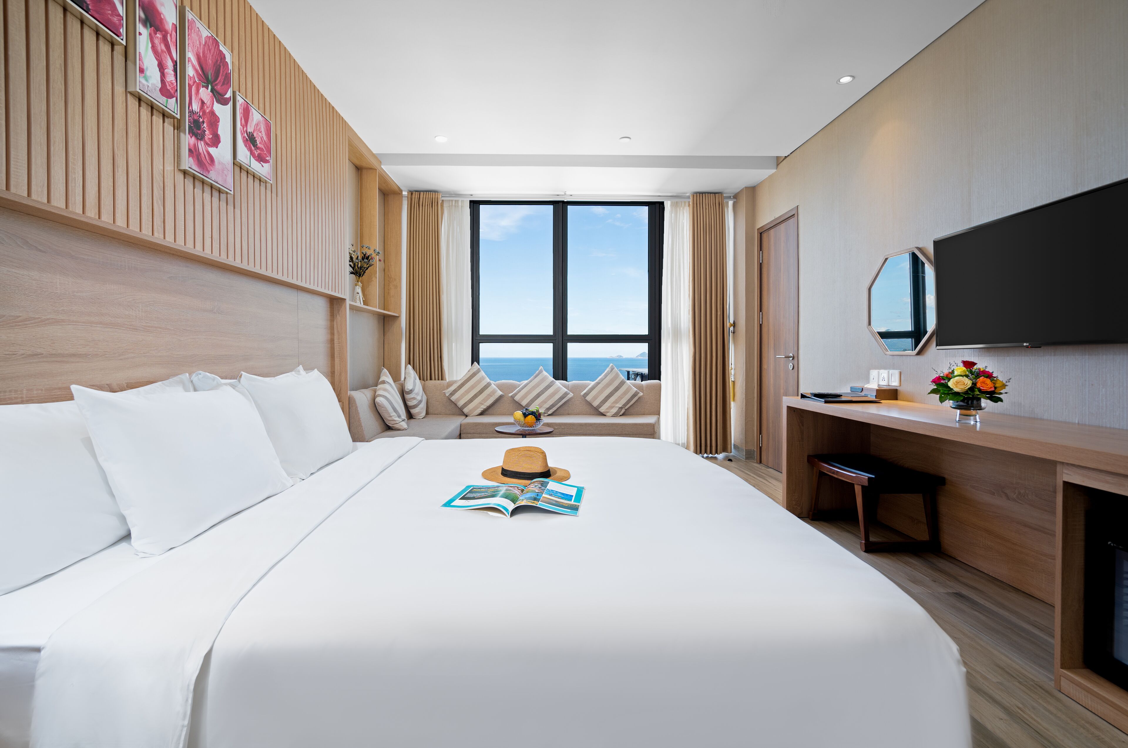 A modern hotel room with a large bed, a desk, a TV, and a view of the sea.