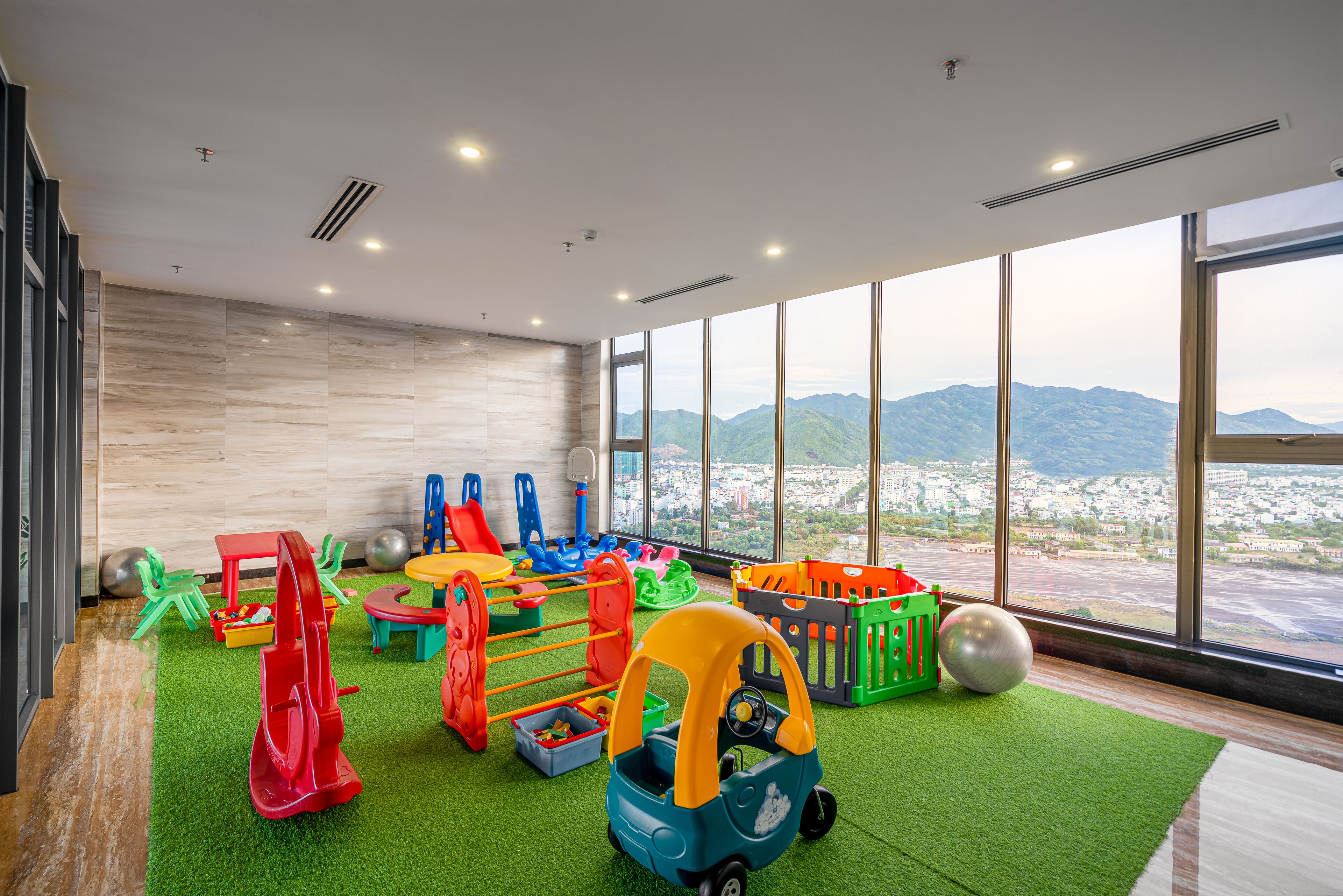 A children's play area with various toys and equipment, large windows with a view of mountains, and a green carpet.