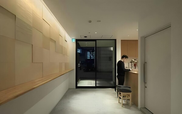 A hallway with a wooden wall, a glass door leading to a dark interior, and a person standing by a counter with a stool.