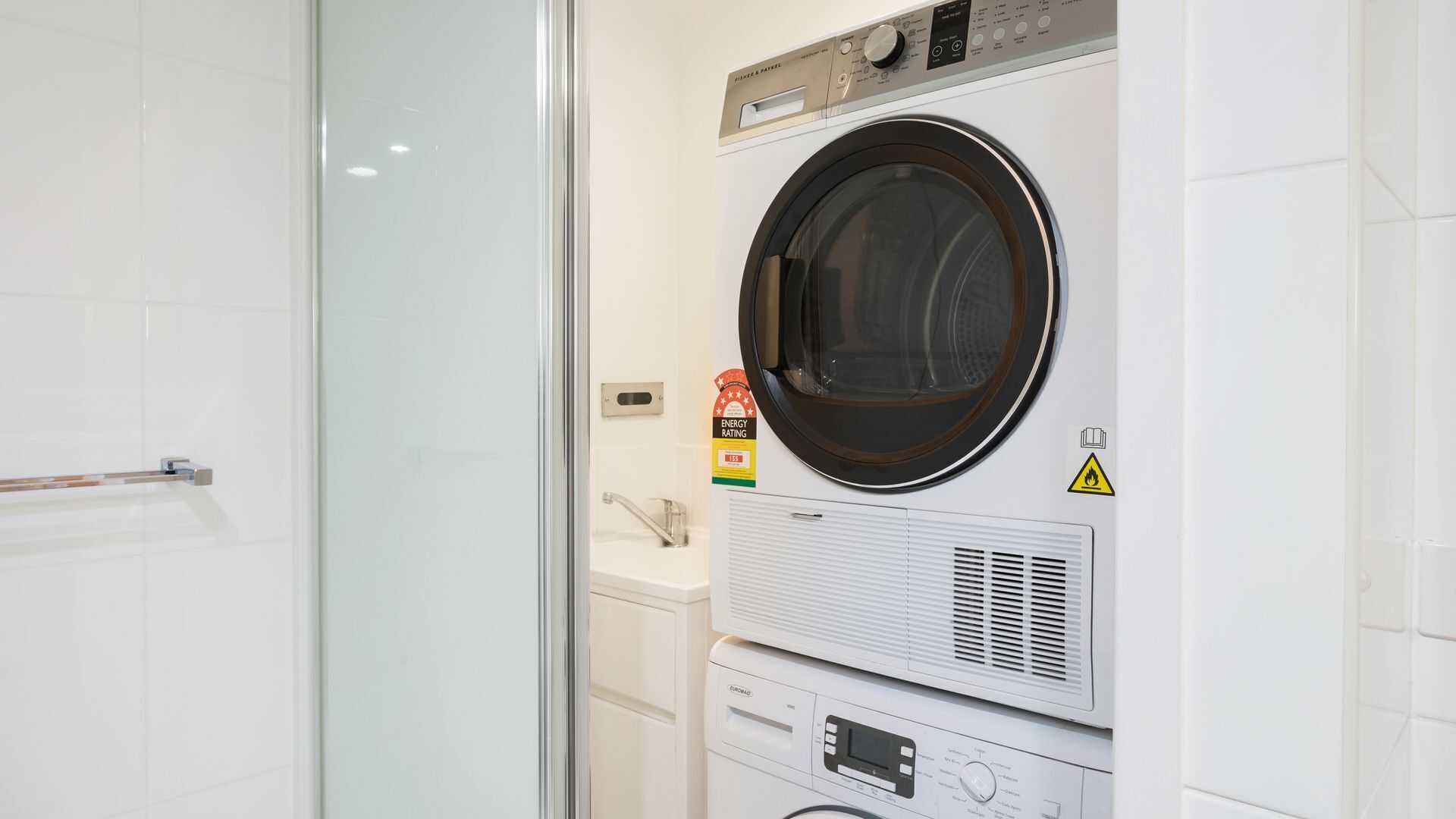 A laundry room with a front-loading washer and dryer, a safety warning sign, and an energy efficiency label.