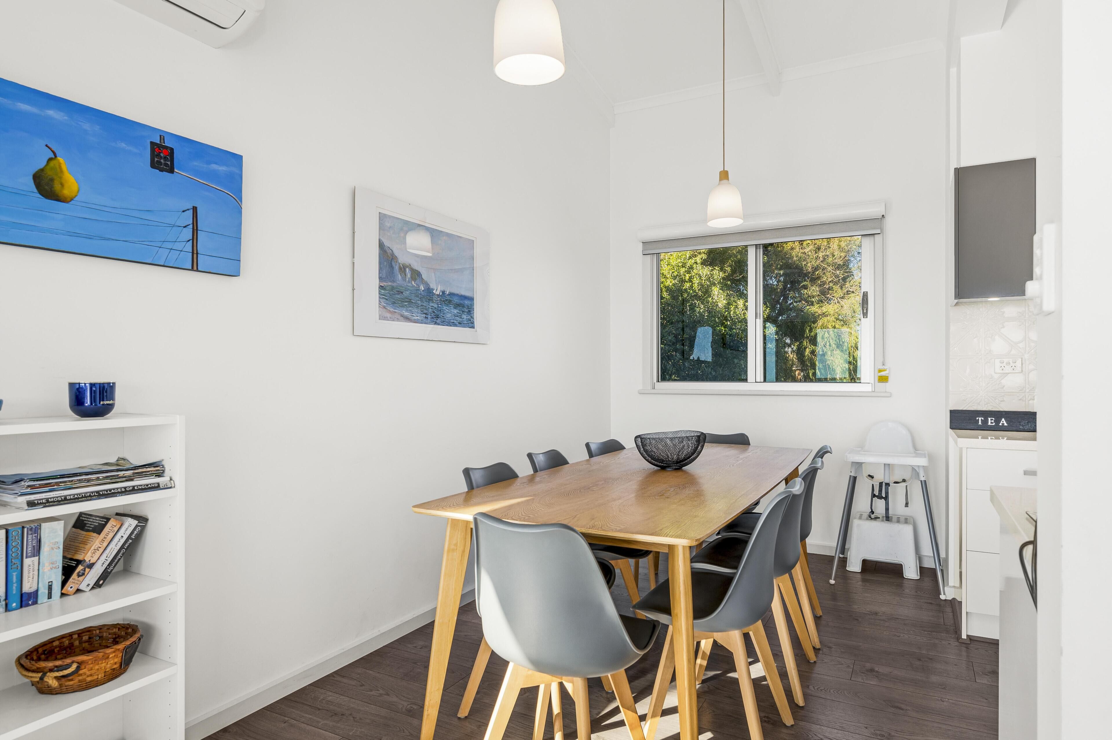 A dining area with a wooden table, grey chairs, a white bookshelf, and a window with a view of greenery.
