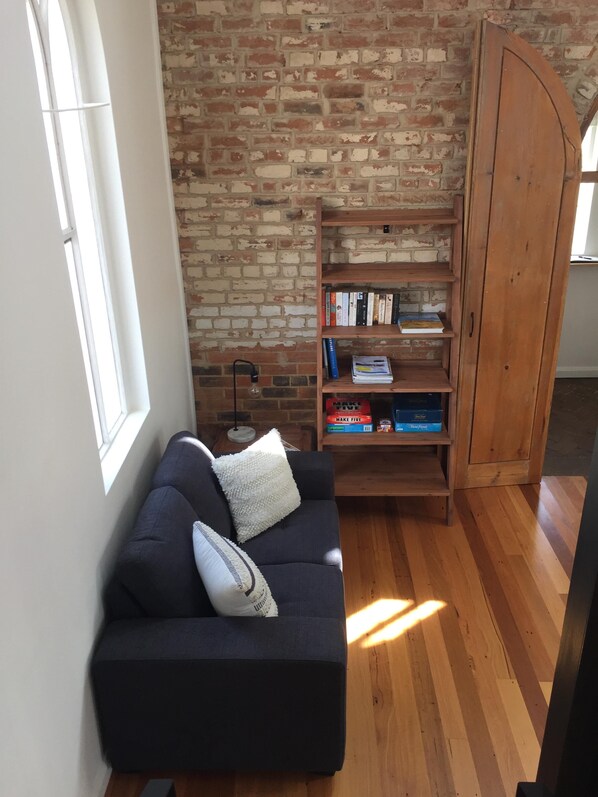 Una habitación con pared de ladrillo, una estantería de madera, un sofá azul oscuro con un cojín blanco y suelo de madera.