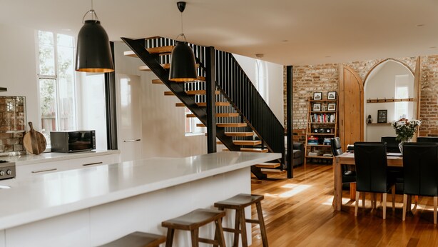 Une cuisine moderne avec un îlot central blanc, des tabourets en bois, un escalier noir et un coin repas comprenant une table et des chaises en bois.