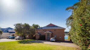 A brick building with a blue roof, a driveway, and a parking area with cars. - Grand Pacific 2 Unit 2 - Ground Floor (NAROOMA)