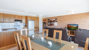 A modern kitchen with wooden cabinets, a white refrigerator, and a dining area with a glass table and chairs. - Grand Pacific 2 Unit 2 - Ground Floor (NAROOMA)