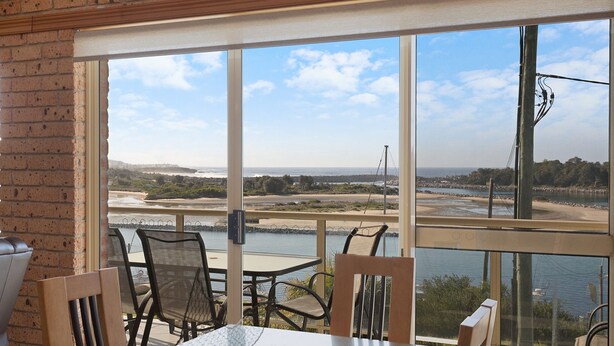 A room with a large glass window overlooking a beach and river. There are chairs and a table inside. - Grand Pacific 2 Unit 2 - Ground Floor (NAROOMA)