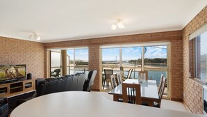 A spacious room with a large dining table, comfortable seating, a television, and large windows offering a view of the beach and ocean. - Grand Pacific 2 Unit 2 - Ground Floor (NAROOMA)
