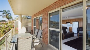 A balcony with a glass door leading to a kitchen area, a dining table with chairs, and a view of palm trees and hills. - Grand Pacific 2 Unit 2 - Ground Floor (NAROOMA)