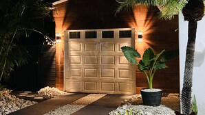 A well-lit garage door with a decorative light fixture above it, surrounded by a gravel pathway and potted plants. - Costa Azul Suite  (Aguadilla)