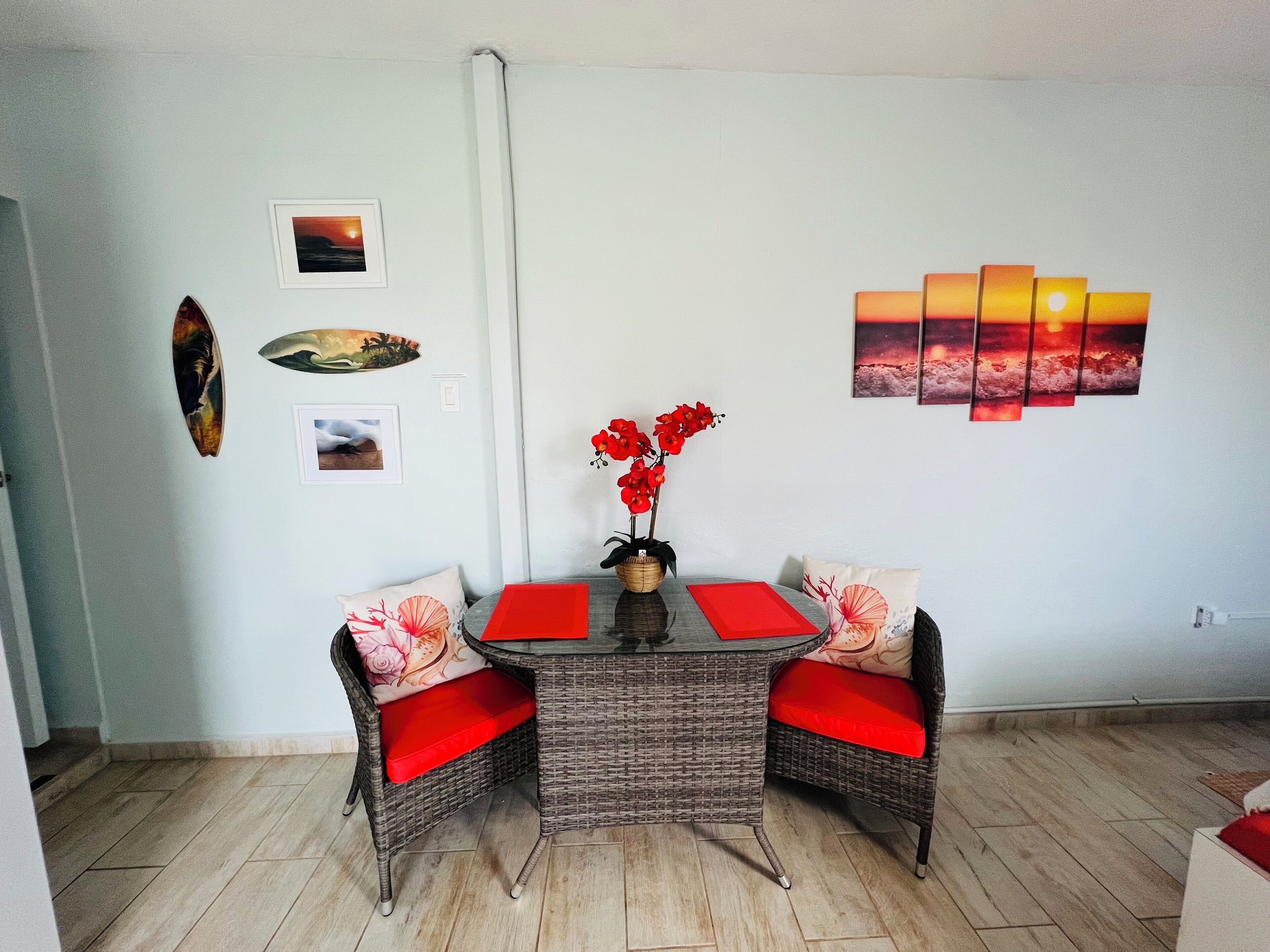 Un área de comedor con sillas de mimbre y cojines rojos, una mesa con camino de mesa rojo y decoración en la pared que incluye un atardecer y un pez.