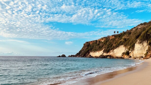 A beach with clear skies, calm waters, and a rocky cliff with vegetation. - Costa Azul Suite  (Aguadilla)