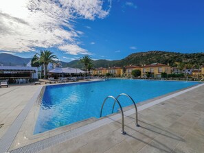 A swimming pool with sun loungers and umbrellas, surrounded by buildings and palm trees.