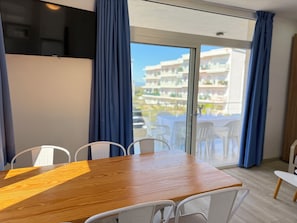 A room with a wooden dining table, white chairs, a TV mounted on the wall, and a view of a balcony with outdoor seating.
