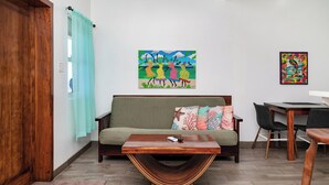 Ein Wohnzimmer mit einem Holztisch, einer Couch mit bunten Kissen, einem Esstisch mit Stühlen und zwei Wandbildern.