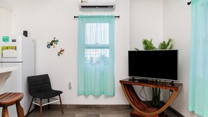 Ein Wohnzimmer mit Fernseher auf einem Holzboden, einem schwarzen Sessel, einem kleinen Tisch und einem Fenster mit hellblauer Gardine.