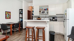 Eine moderne Küche mit weißen Schränken, einer Mikrowelle, einer Kaffeemaschine und einem Kühlschrank. Zwei hölzerne Barhocker und ein Mülleimer sind ebenfalls vorhanden.
