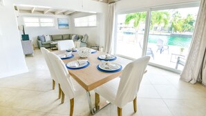 A dining area with a wooden table set for four, white chairs, and a view of a swimming pool through large windows. - WATERFRONT Key Colony Beach Retreat 37’ Dock Cabana Club- Kayak & Bikes- Parking (Marathon)