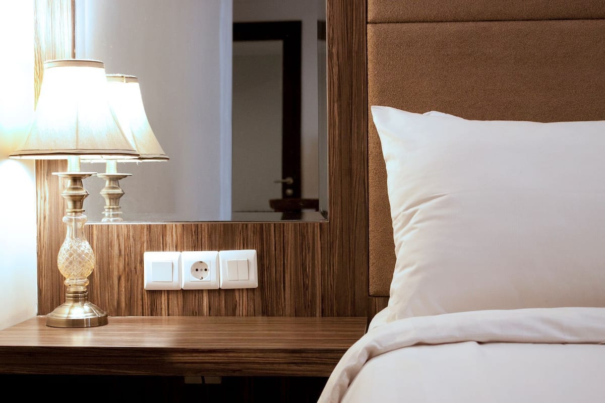 Una cama bien tendida con un almohadón blanco y una lámpara sobre una mesita de noche de madera. Hay tres interruptores en la mesita.