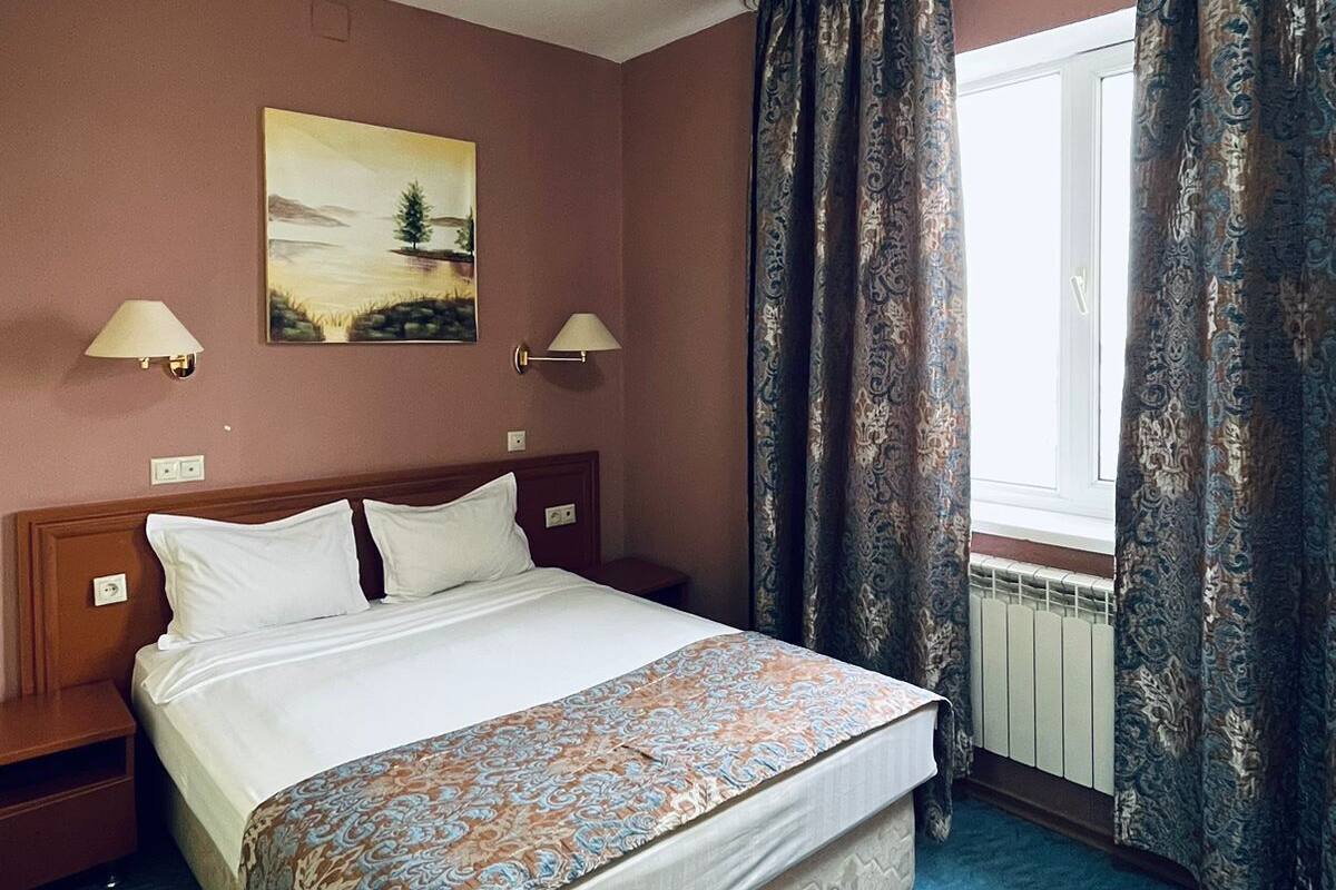 A hotel room with a large bed, two bedside lamps, a painting on the wall, and a window with curtains.