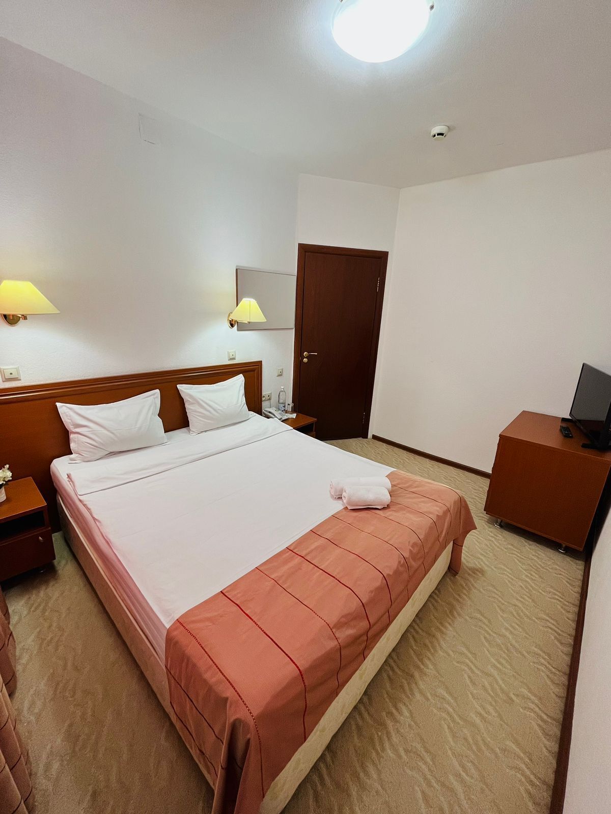 Ein Hotelzimmer mit Bett, zwei Nachttischlampen, einem Holz-Kopfteil, einer Tür, einem Nachttisch, einem Fernseher und einem kleinen Schrank.
