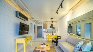 Uma sala de estar moderna com um sofá, mesa de centro e cadeiras amarelas. Ao fundo, há uma área de cozinha com uma mesa de jantar e cadeiras.