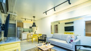 Uma sala de estar moderna com sofá, mesa de centro e cadeiras. Há uma área de cozinha com mesa e cadeiras. Uma televisão de tela plana está fixada na parede.