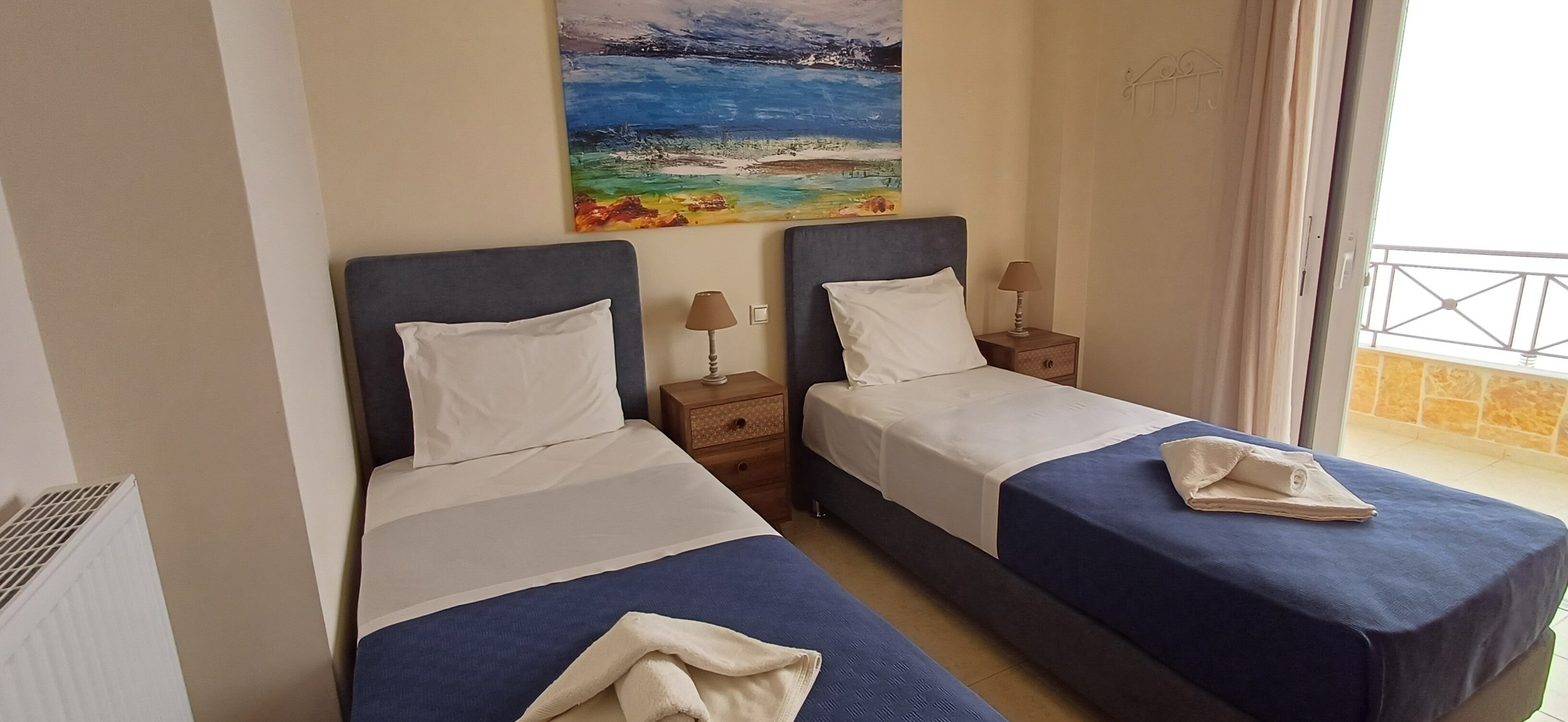 A hotel room with two beds, each with a blue bedspread and white pillows. There is a painting of a beach scene on the wall above the beds.