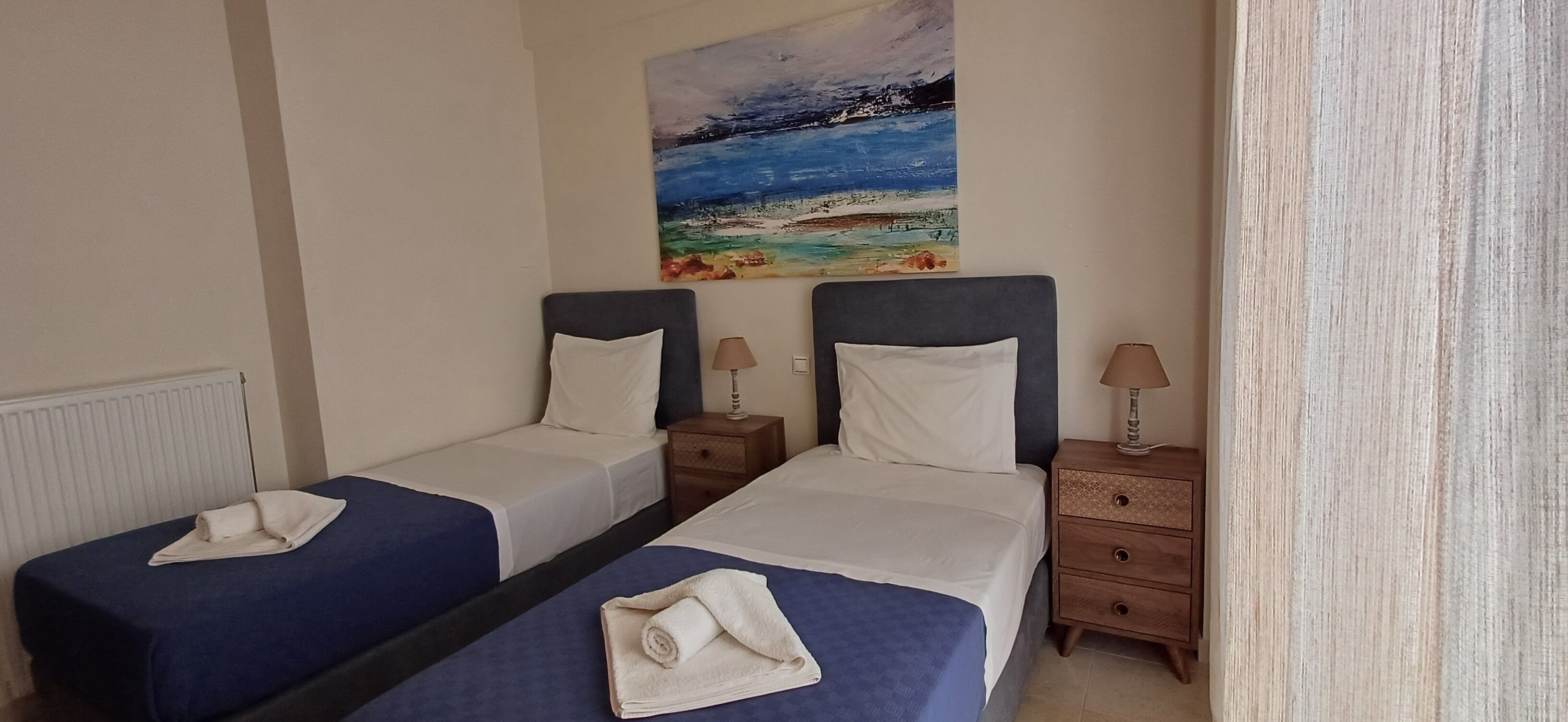 Habitación de hotel con dos camas individuales, cada una con cubrecama azul y almohadas blancas, una mesita de noche con lámpara y un cuadro con una escena de playa en la pared.