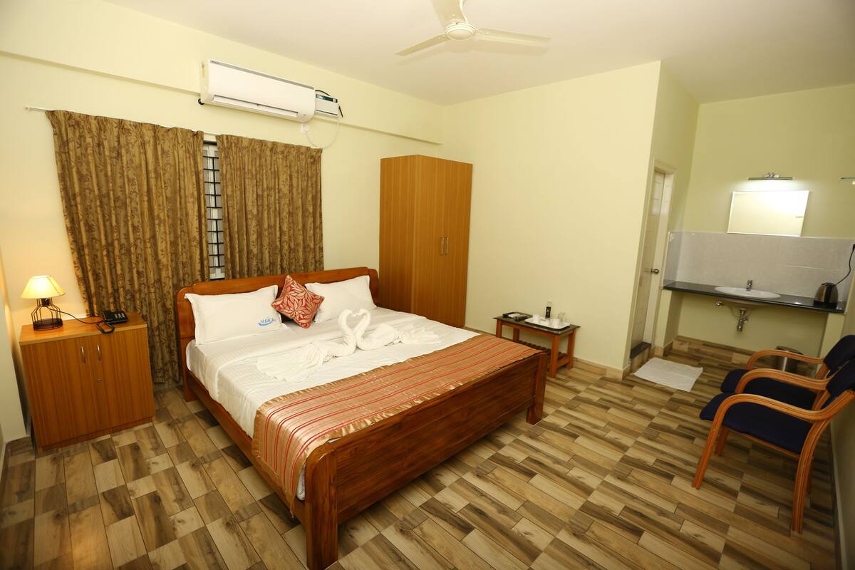 Habitación de hotel con cama de madera, armario, escritorio con lámpara, silla y baño con espejo y lavamanos.