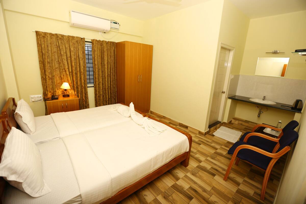 Habitación de hotel con cama, armario, escritorio con lámpara, silla, baño con lavamanos y espejo.