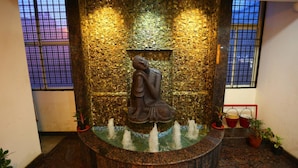 A stone fountain with a seated figure, surrounded by water jets, set against a textured wall with windows. - Hotel The Shack (Bengaluru)