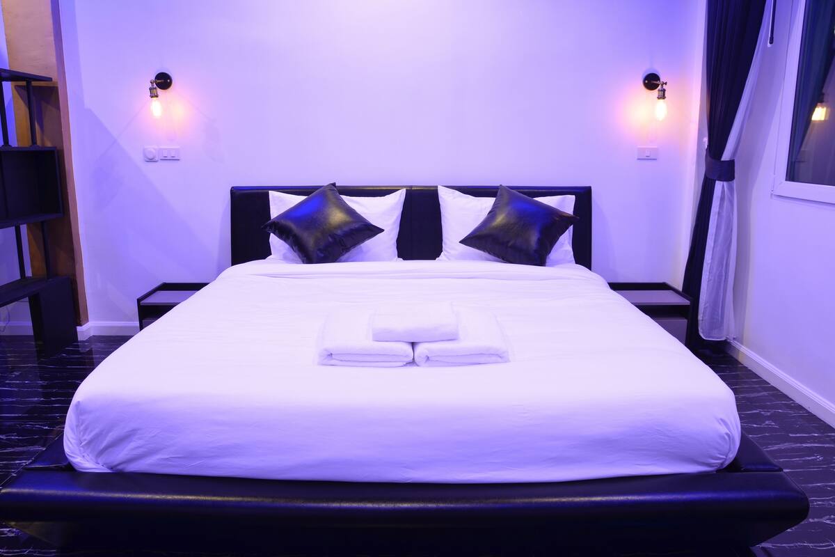 Ein ordentlich gemachtes Bett mit weißen Leinen und zwei schwarzen Kissen, ein Nachttisch und Wandleuchten.