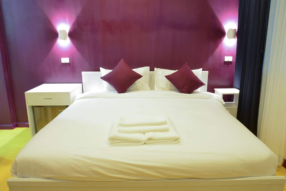 Ein ordentlich bezogenes Bett mit weißen Leinen und einem gefalteten Handtuch, zwei dunkle Kissen und eine violette Akzentwand mit Wandleuchten.
