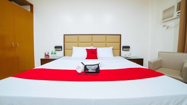Une chambre d’hôtel avec un grand lit, une couvre-lit rouge et blanche, un oreiller rouge, une petite table avec un téléphone et une bouteille, et une chaise.