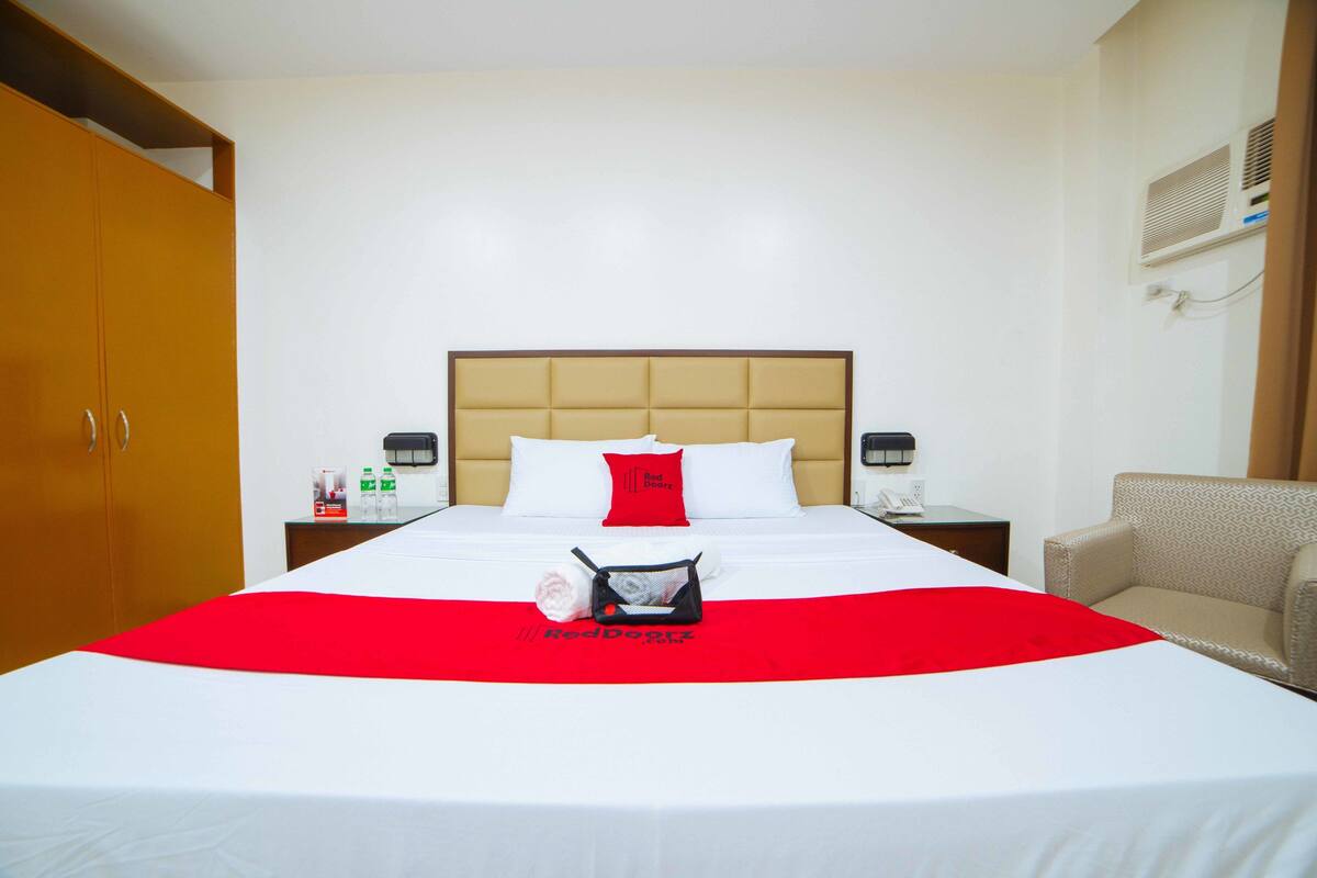 Une chambre d’hôtel avec un grand lit, une couvre-lit rouge et blanche, un oreiller rouge, une petite table avec un téléphone et une bouteille, et une chaise.