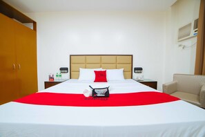 A hotel room with a large bed, a red and white bedspread, a red pillow, a small table with a phone and a bottle, and a chair.