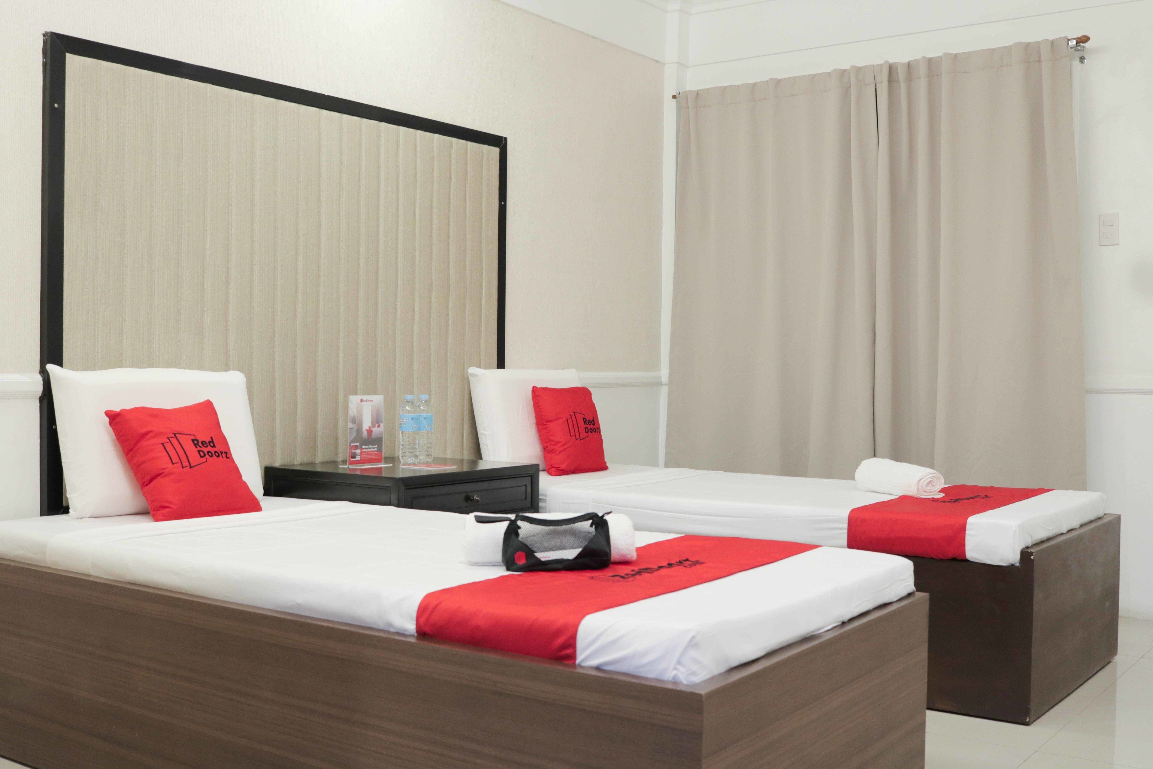 A hotel room with two beds, each with a red blanket and pillow, a wooden nightstand with a water bottle, and a large mirror.