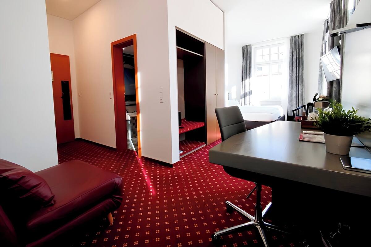 Habitación de hotel con alfombra roja, un escritorio con silla, una planta en maceta y una ventana con cortinas.