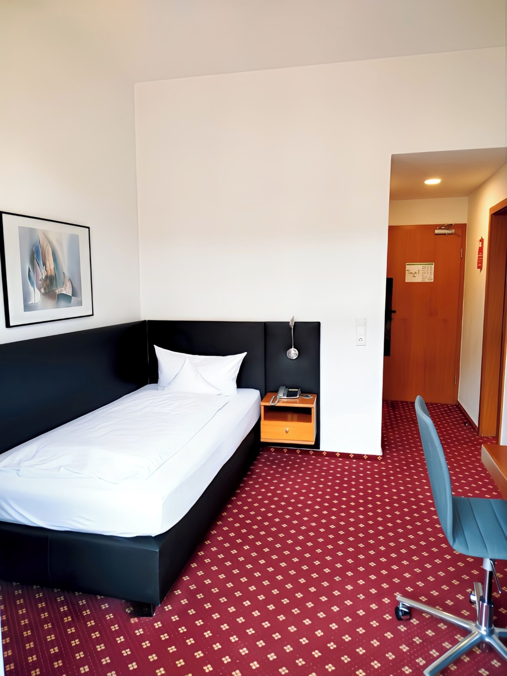 Une chambre d’hôtel avec un lit simple, une table de chevet avec une lampe, une chaise et une moquette à motifs.