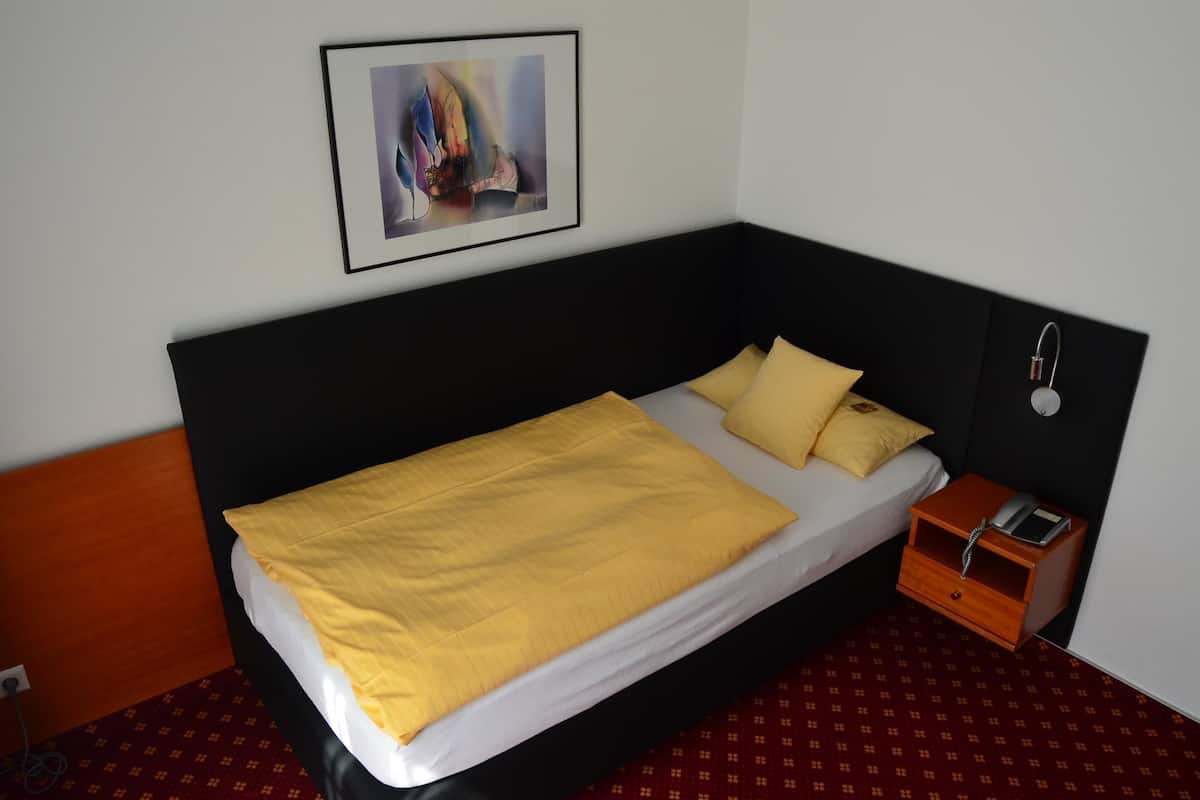 Una cama individual con cabecera negra, una colcha amarilla y una mesita de noche de madera con un teléfono.