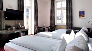A hotel room with two beds, a desk with a flat-screen TV, a chair, a small table, and a window with curtains. - Hotel Schwert (Rastatt)