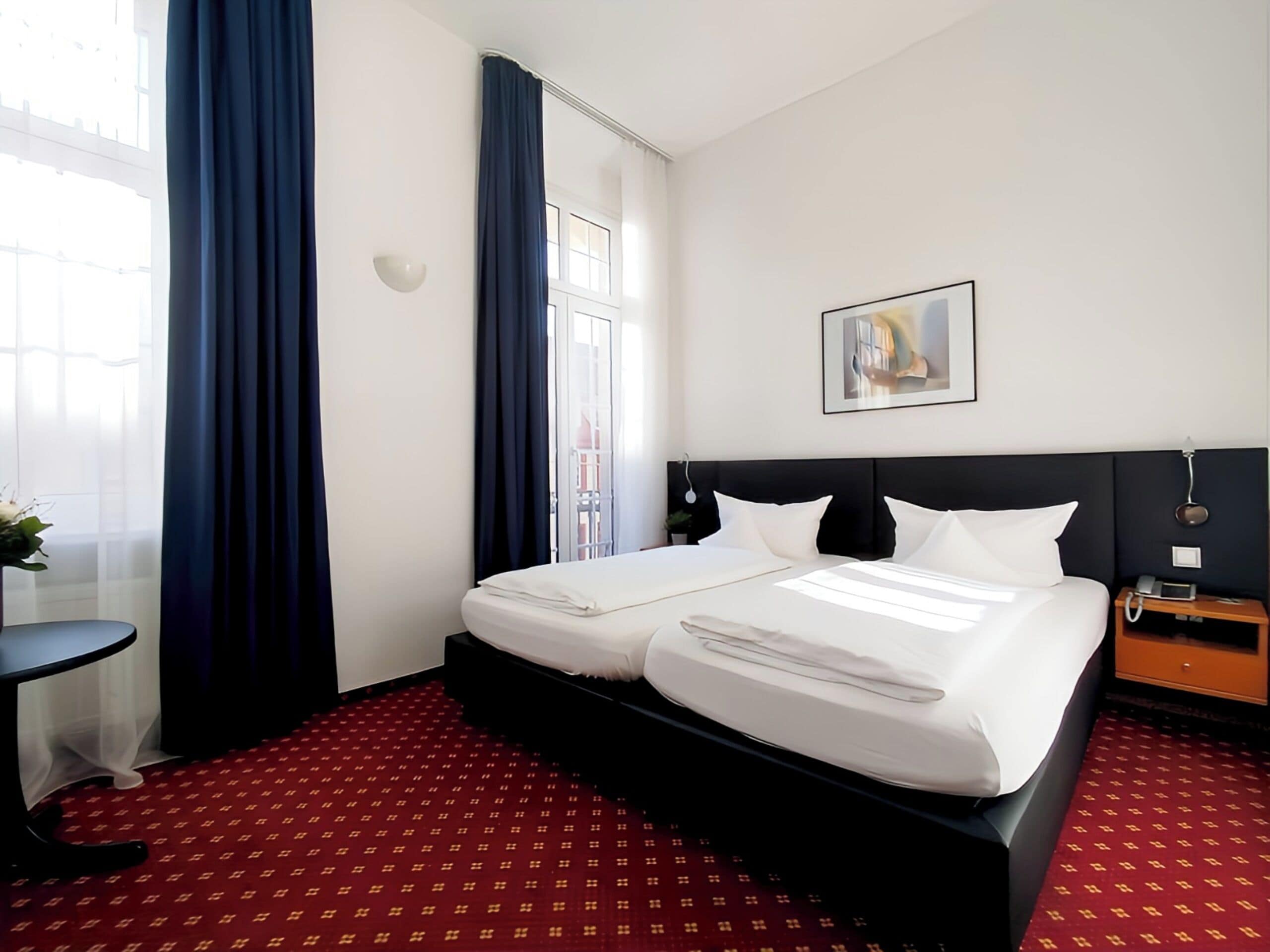 A hotel room with a double bed, a small bedside table, a nightstand with a phone, and a window with curtains.