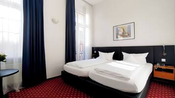 A hotel room with a double bed, a small bedside table, a nightstand with a phone, and a window with curtains.